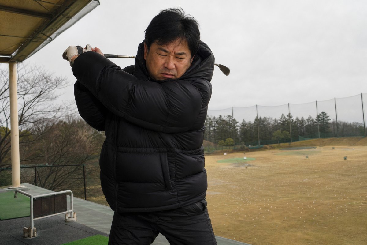 厚手の上着を着込んで窮屈そうにスイングする日本人男性ゴルファー